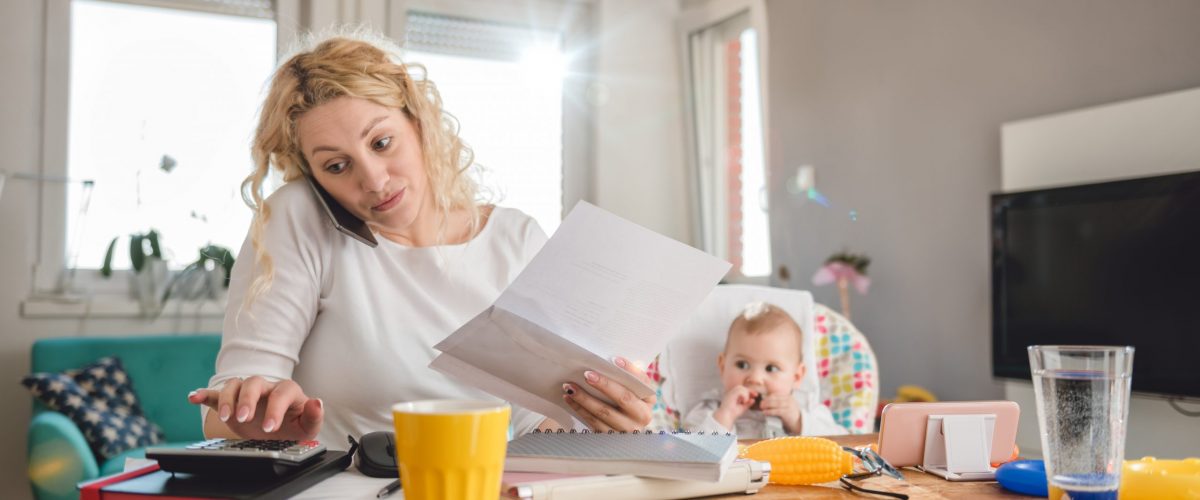 Mother holding letter, using calculator, talking on smart phone at home office and taking care of her baby